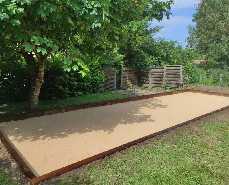 Terrain de pétanque bordé de bois sous des arbres, sol sableux stabilisé