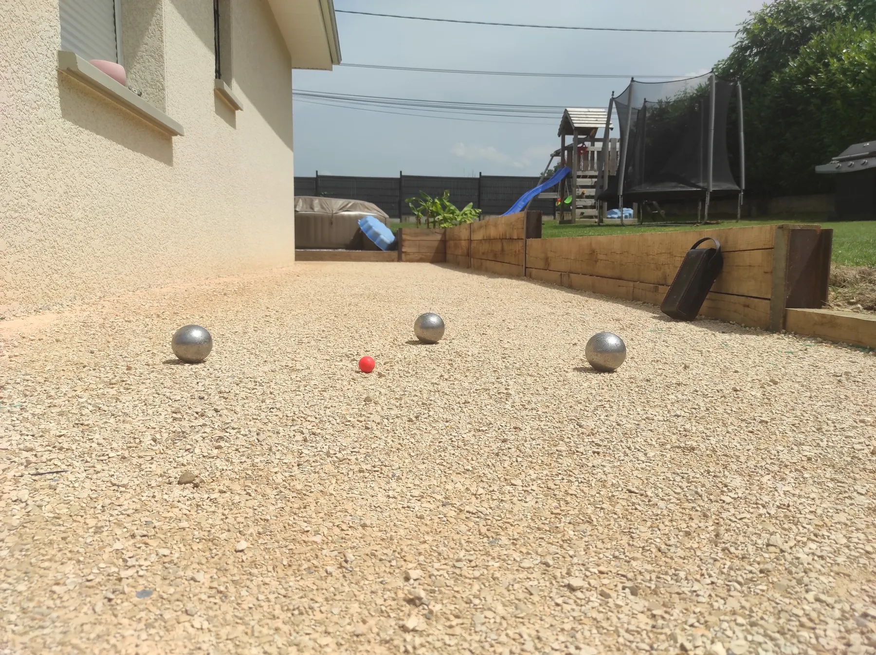 Terrain de pétanque en gravier clair avec boules, aire de jeux en arrière-plan