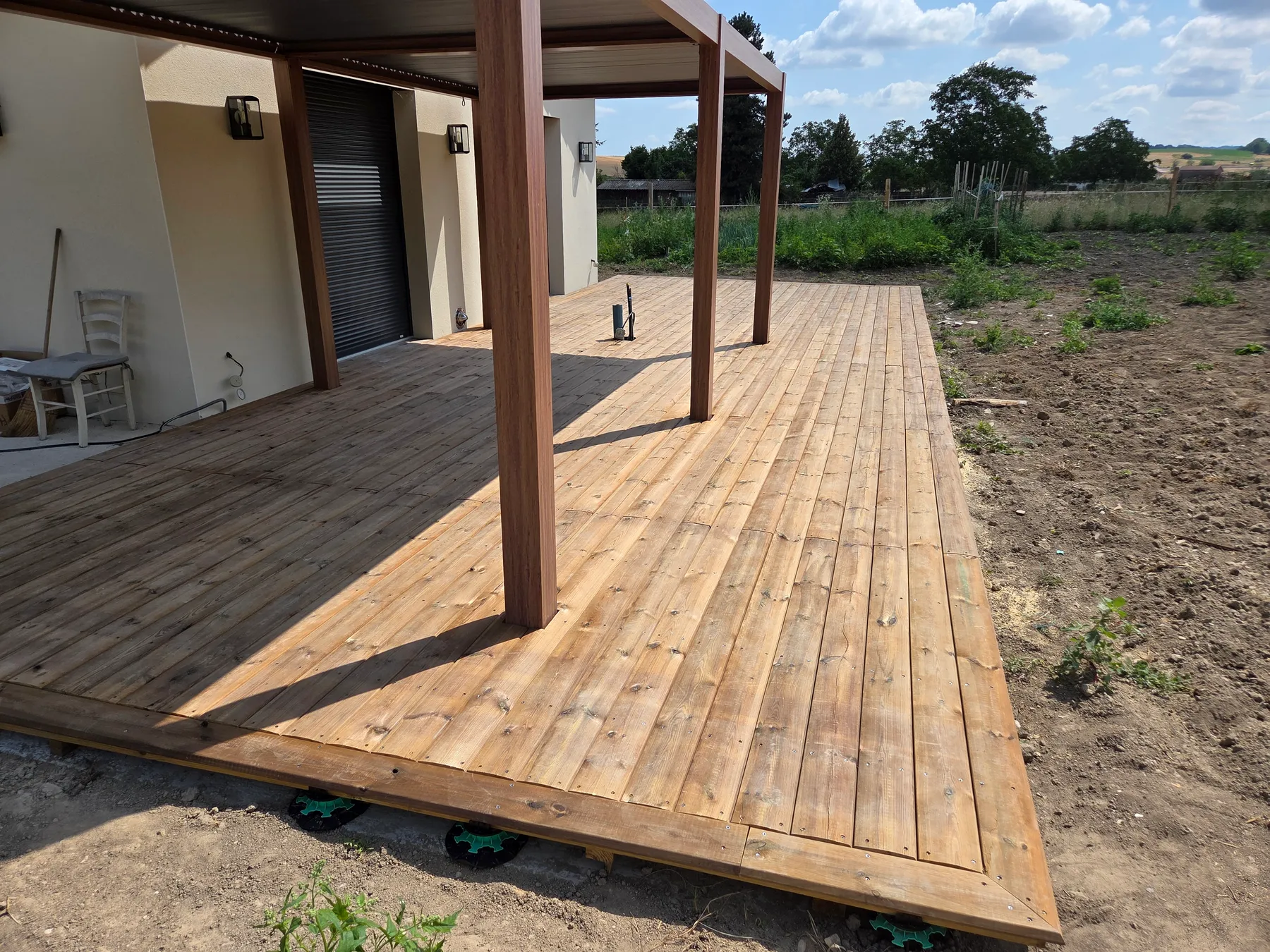 Platelage en lames de bois posé sur plots réglables sous une pergola en aluminium, contre le mur d’une maison neuve